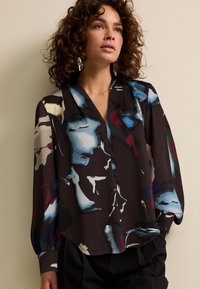 Donkere blouse met een V-hals, voorzien van een abstract multicolor patroon in blauw, bordeauxrood en crème. Lange mouwen met elastische manchetten.