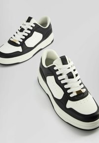 Black and white sneakers with a smooth leather upper, textured accents, white laces, and a gold eyelet detail at the tongue.