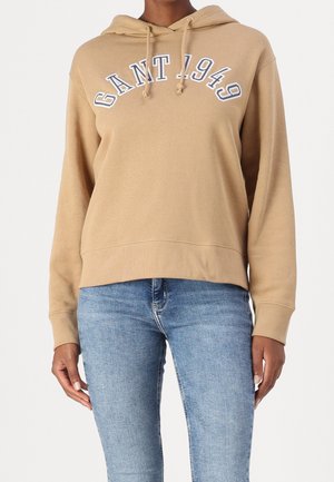 Persoon draagt een beige GANT-hoodie met de tekst "GANT 1949" en lichtblauwe jeans, staand tegen een effen achtergrond.