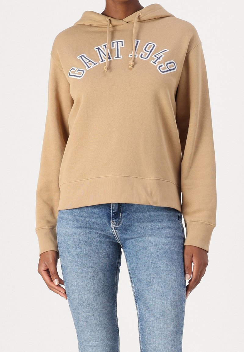 Personne portant un hoodie GANT beige avec le texte "GANT 1949" et un jean bleu clair, debout devant un fond uni.