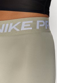 Svijetlozelene tajice s širokim pojasom na kojem se nalazi bijeli natpis "NIKE PRO". Tkanina izgleda glatko i prilagođeno, idealno za sportske aktivnosti.