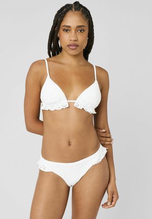 Femme aux cheveux tressés portant un bikini blanc en broderie anglaise avec des bords volantés, debout devant un fond gris uni.