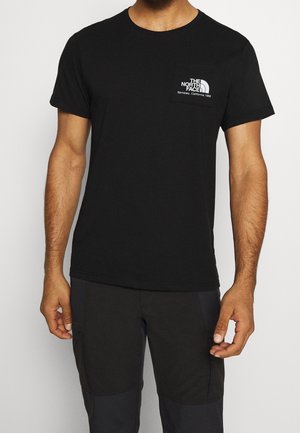 Mężczyzna ubrany w czarną koszulkę z krótkim rękawem z logo i napisem The North Face na lewej stronie klatki piersiowej, w połączeniu z czarnymi spodniami.