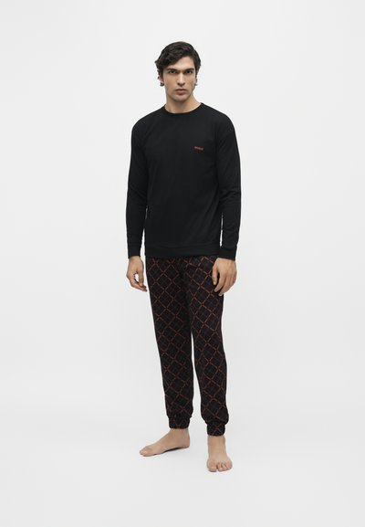 Homme debout pieds nus portant une chemise noire à manches longues et un pantalon noir avec des motifs géométriques rouges sur un fond blanc uni.