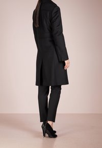 Cappotto nero con un design su misura, maniche lunghe e colletto strutturato. Abbinato a pantaloni neri aderenti e scarpe con tacco.