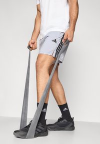 Bandas de resistencia grises sostenidas por una persona que lleva una camiseta blanca, pantalones cortos gris claro con rayas negras y zapatos deportivos negros.