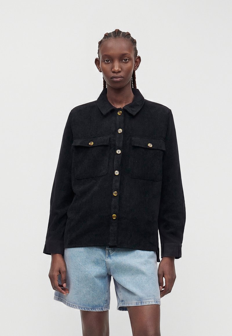 Jonge persoon met gevlochten haar draagt een losse zwarte corduroy jas met knopen over een lichtblauwe denim short.