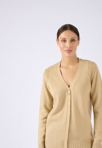 Beiger Cardigan aus Strickstoff mit V-Ausschnitt, Knopfleiste, geripptem Saum und dezenten strukturierten Akzenten an den Schulternähten.