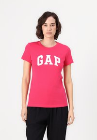 Lyserød bomulds t-shirt med et stort hvidt "GAP" logo i fed skrift, rund halsudskæring og korte ærmer, præsenteret mod en hvid baggrund.