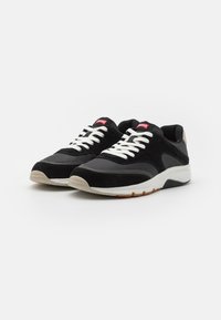 Schwarze Sneakers aus Wildleder und Mesh-Material, mit weißen Schnürsenkeln und einer gepolsterten Sohle. Rotes Markenetikett auf der Zunge. Leichtes Design.