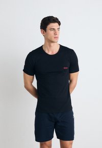 Svart t-skjorte med korte ermer og rund hals, med en liten rød "HUGO"-logo på venstre bryst; satt sammen med marineblå shorts.