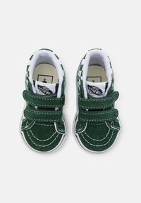 Zapatillas de terciopelo verde con costuras blancas y correas de Velcro. Patrón de tablero de ajedrez en la parte superior. Suelas de goma con punta redonda.