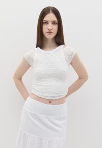 Top bianco corto con texture in pizzo floreale e scollatura rotonda. Abbinato a una gonna bianca a balze, creando un look leggero e stratificato.