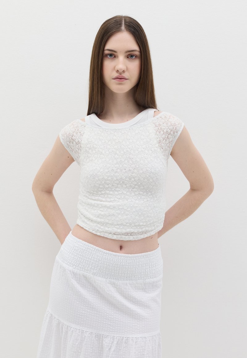 Top bianco corto con texture in pizzo floreale e scollatura rotonda. Abbinato a una gonna bianca a balze, creando un look leggero e stratificato.