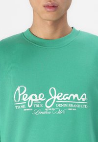 Sudadera de algodón verde con cuello redondo acanalado y logotipo impreso en blanco. Presenta tipografía elegante y detalles de acento.