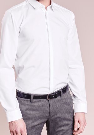 Mężczyzna w białej koszuli zapinanej na guziki, wpuszczonej w szare, teksturowane spodnie z czarnym skórzanym paskiem na tle o neutralnym kolorze.