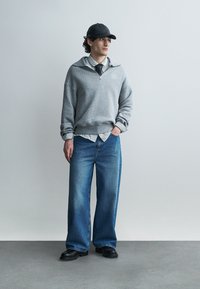 Νέος άνδρας που φοράει μαύρο καπέλο, γκρι ζακέτα με φερμουάρ πάνω από άσπρο πουκάμισο με μαύρη γραβάτα, φαρδιά μπλε τζιν και μαύρα παπούτσια, στέκεται μπροστά σε γκρίζο τοίχο.