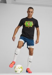 T-shirt nera con grafica colorata "Forever Brasileiro", pantaloni corti blu con accenti geometrici, calzini bianchi e scarpini da calcio rosa acceso.