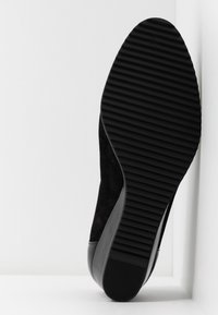 Semelle de chaussure noire avec motif de rainures horizontales, placée inclinée contre un coin blanc.