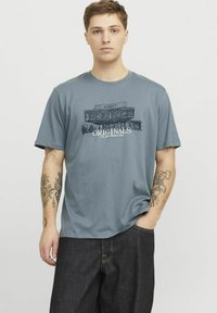 Tricou din bumbac gri cu un design grafic ce include textul "ORIGINALS" în alb, mâneci scurte și o croială lejeră.