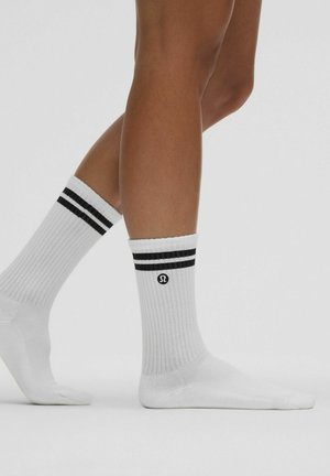 Valkoiset ribatut sukat, joissa on mustat raidat resorissa ja logo nilkassa. Pehmeä rakenne, puoliväliin säärtä ulottuva pituus ja istuva muotoilu.