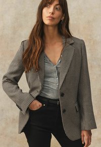 Houndstooth mønsteret blazer i sort og creme, med notched lapels, to knapper og frontlommer. Parret med en grå V-udskæring top.