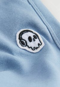 Helles blaues Gewebe mit einem weißen gestickten Patch, der ein schwarzes Totenkopfdesign und Kopfhörer zeigt, hebt die Textur und Details hervor.