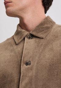 Katoenen overhemd in lichtbruin, met een kraag, knoopsluiting en een textuur met verticale ribbels. Twee zwarte knopen zichtbaar.