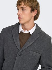 Manteau en laine gris charbon avec col cranté, fermeture à un bouton et une poche poitrine. Porté sur un pull marron.
