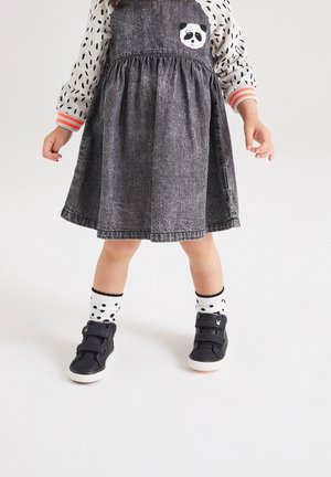 Tout-petit portant une robe grise avec un patch panda, une chemise à manches longues à pois, des chaussettes blanches à pois et des chaussures noires à scratch, debout sur un sol blanc.