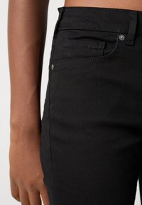 Acercamiento de una persona que lleva puestos unos pantalones de mezclilla negra, con un bolsillo frontal visible y detalles de remaches en el área de la cadera.