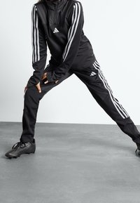 Czarny sportowy dres z białym wzorem trzech pasków na rękawach i nogawkach. Posiada półzamknięty kołnierz i dopasowany krój. Czarne korki.