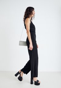 Femme aux cheveux bouclés portant une combinaison noire sans manches, des talons compensés noirs et un petit sac à main en argent marchant contre un fond blanc.