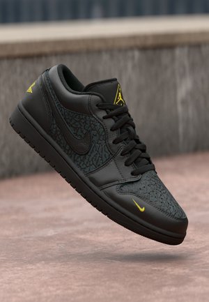 Sorte og mørkegrå Nike Air Jordan sneakers med gule logoer og teksturerede mønstre, svævende over en tekstureret betonoverflade.