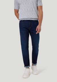 Man in opgerolde donkerblauwe jeans, witte sneakers en een lichtgrijs gemêleerd shirt met korte mouwen, staand met één hand in zijn zak.