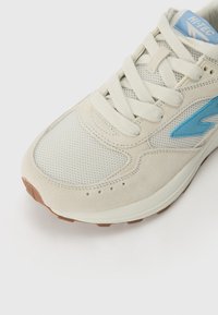 White sneaker featuring a mesh upper, suede accents, light blue branding, and a brown rubber outsole. Close-up angle highlighting details.