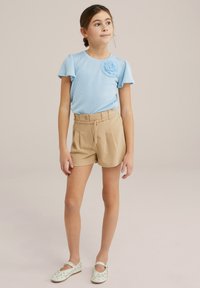 Tricou cu maneci scurte, de culoare albastră deschisă, cu detaliu floral, asociat cu pantaloni scurți bej, cu talie înaltă, și încălțări Mary Jane albe cu accente verzi.