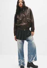 Giacca bomber di pelle marrone con due tasche con zip, gonna nera plissettata, jeans larghi azzurri chiari con macchie bianche consumate e stivali neri alla caviglia.