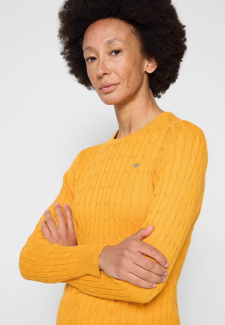 Żółty sweter robiony na drutach o prążkowanej strukturze, z okrągłym dekoltem i wzorem warkoczowym; zawiera małe haftowane logo na klatce piersiowej.