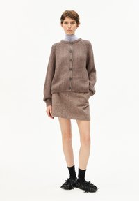 Frau in einem braunen, geknöpften Strickcardigan, passendem Rock, Lavendelrollkragenpullover, schwarzen Socken und schwarzen Quasten-Loafers, die vor einem weißen Hintergrund steht.