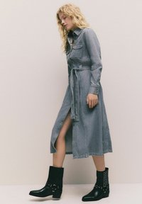 Frau mit lockigem blonden Haar, die geht, trägt ein graues Denim-Kleid mit Gürtel, einem Schlitz vorne und schwarzen Stiefeletten mit silbernen Nieten.