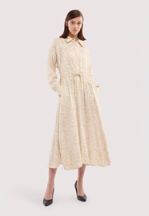 Kvinde står med hænderne i lommerne iført en lang beige mønstret kjole med krave og bånd i taljen, kombineret med sorte hæle.