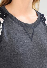 Sweatshirt gris clair avec des accents tissés texturés sur les épaules. Comprend un col rond et des manches raglan.