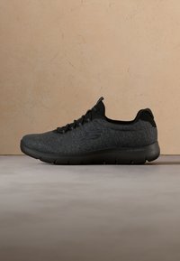 Chaussure de sport légère Skechers gris foncé avec semelle noire et logo discret sur le côté, présentée sur une surface lisse sur fond beige.