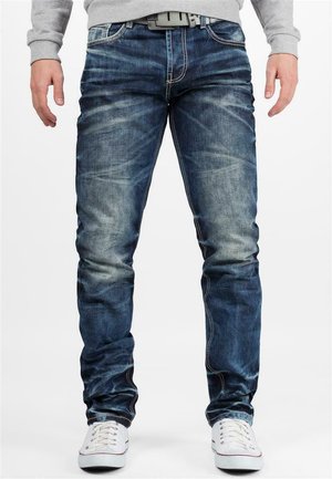 Denim jeans in dark blue with fading effects, slim fit design, visible stitching, and five-pocket styling. Worn with white sneakers.