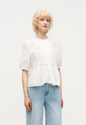 Femme aux cheveux blonds courts et bouclés portant un chemisier blanc à motif de dentelle et un jean bleu clair à taille haute, debout devant un fond uni.