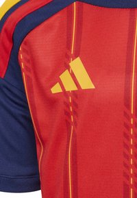 Ružová a námornícka športová dresová rohová časť so žltým logom Adidas a tenkými vertikálnymi žltými pruhmi na červenej látke.