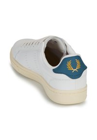 Zapatilla blanca con parte superior de cuero suave, suela de goma beige y un tirador del talón en color azul verdoso con diseño de hojas doradas. Cierre con cordones y líneas de ventilación.
