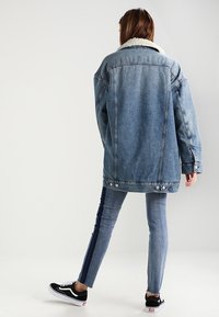 Džínová bunda s beránkovým límečkem, dlouhými rukávy a detailem na zadní kapse, spárovaná s úzkými džíny s tmavými postranními pruhy a černými teniskami.