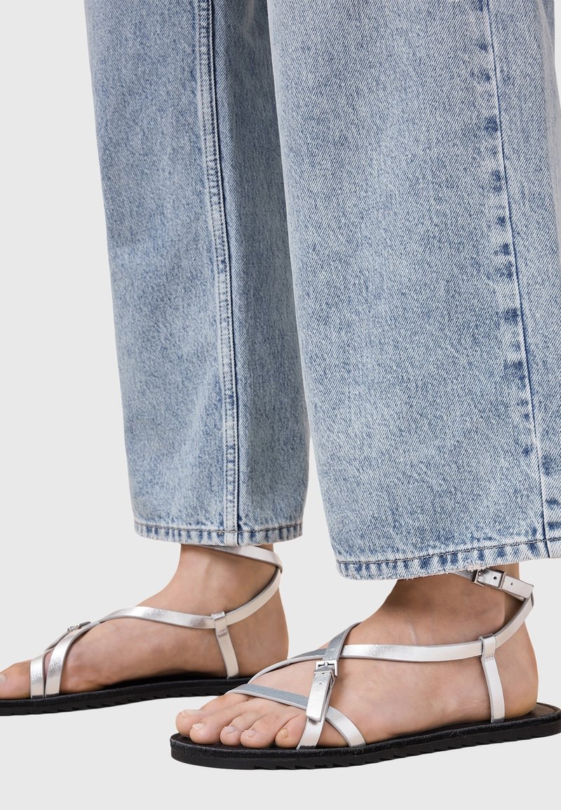 Pieds portant des sandales plates argentées à brides, associées à un jean en denim bleu clair à jambes larges sur un fond uni.